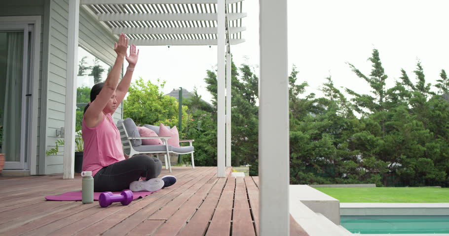 Mature woman in pink top finishing weights on deck, moving into meditation for cooldown, copy space. Mindfulness, yoga, relaxation, serenity, stretch, patio, poolside