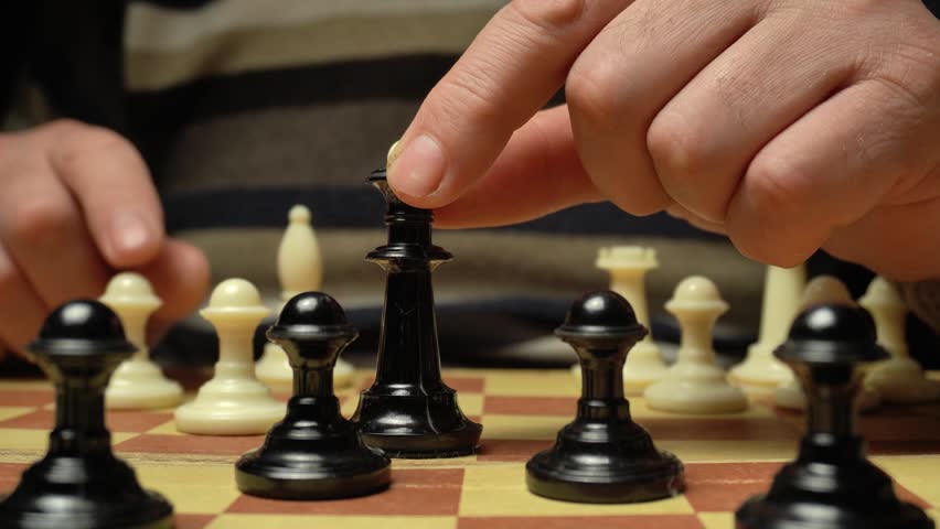 Close-up of a hand positioned above a black chess piece on a wooden chessboard, showcasing the movement of the piece while white pawns and other pieces are visible in the background