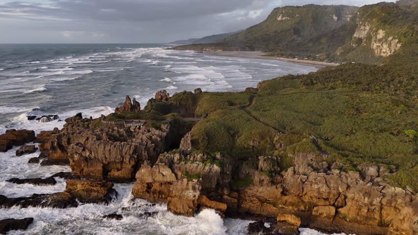 Panoramic aerial view of Pancake Rocks in Punakaiki on West Coast of New Zealand’s South Island at sunset, waves crashing against rugged cliffs under warm golden light. Holiday trip.