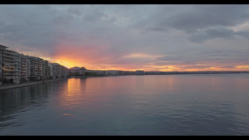 Cinematic aerial tracking shot over the sea looking at the Thessaloniki city waterfront during a spectacular colorful sunset, Greece. Water reflections and clouds.