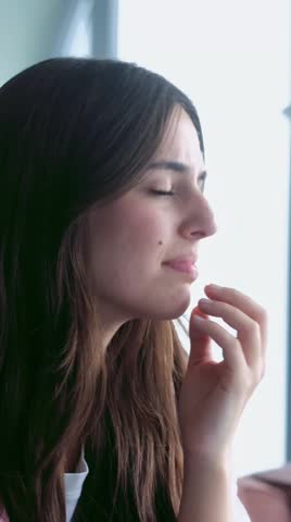 A beautiful young woman in deep thought transitions from a subtle sadness to a hopeful gaze, reflecting on her emotions by a bright window. Her pensive expression conveys inner strength and quiet contemplation.