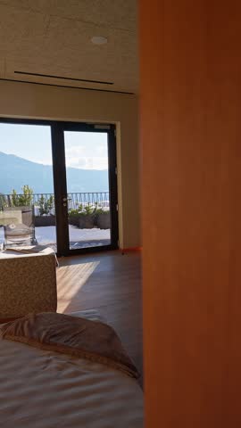 A barefoot guest in a white robe steps toward tall glass doors, a snow rimmed balcony, and wide mountain ridgelines in a wood paneled spa suite with warm LED lighting.