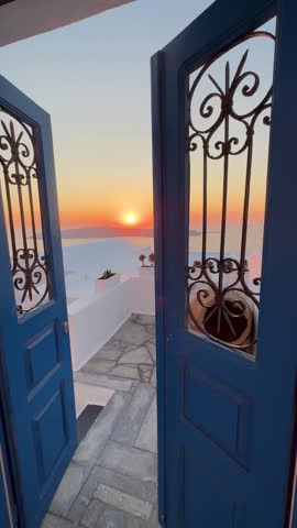 Beautiful sunset in Santorini island, Greece. Passing through the blue door to the terrace. White architecture and sea view at sunset. Travel and summer vacation concept. Vertical video
