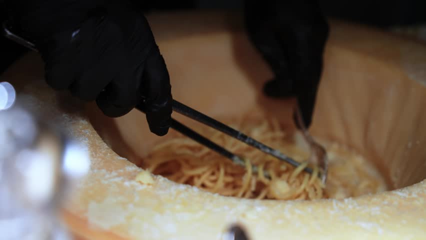 Close up shows gloved hands twirling spaghetti in a Parmesan wheel, scraping to coat pasta in melted cheese. Warm tones and shallow depth of field isolate action.