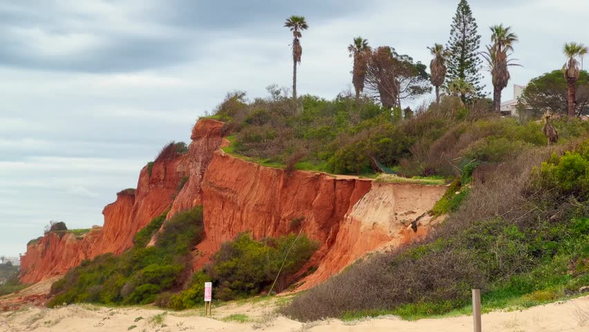 View of empty Praia beach with steep rock cliff, ocean waves, stones, wet golden sand and green vegetation at wild coast, Portugal.