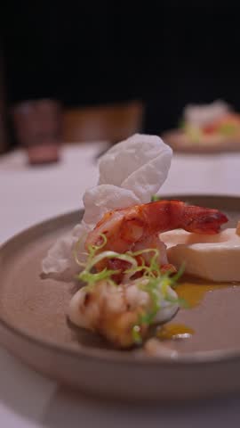 Tight vertical view shows char grilled prawn on white mousse, seared bites, puffed crisps, microgreens, and golden sauce on stoneware, slow tilt in warm light.