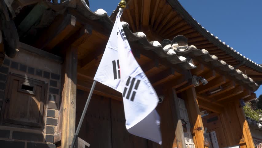 South Korean national flag Taegukgi waving on a pole in front of a traditional Korean hanok wooden architecture with tiled roof under a clear blue sky in Bukchon Hanok Village. Seoul, South Korea