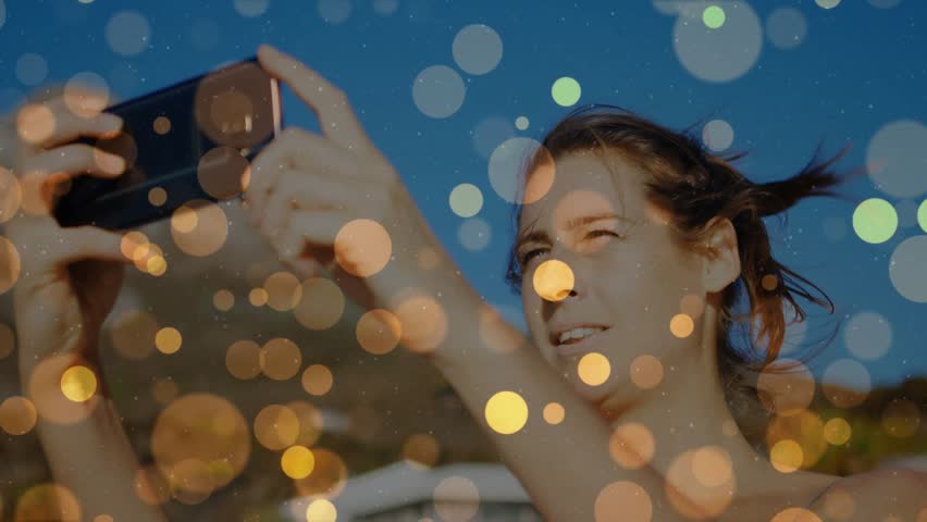 Woman raising smartphone, framing travel shot to capture coast while bokeh drifting across frame. Outdoors, seaside, coastal, sunlit, phone, casual, traveler