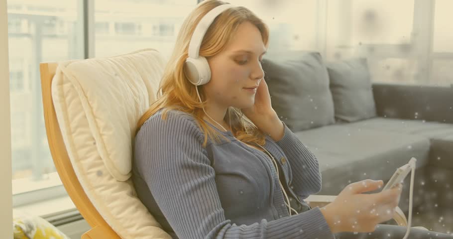 Woman adjusting headset, tapping phone near window, playing audio with particles floating for music. Headphones, tablet, daylight, cozy, cityscape, livingroom, relaxed