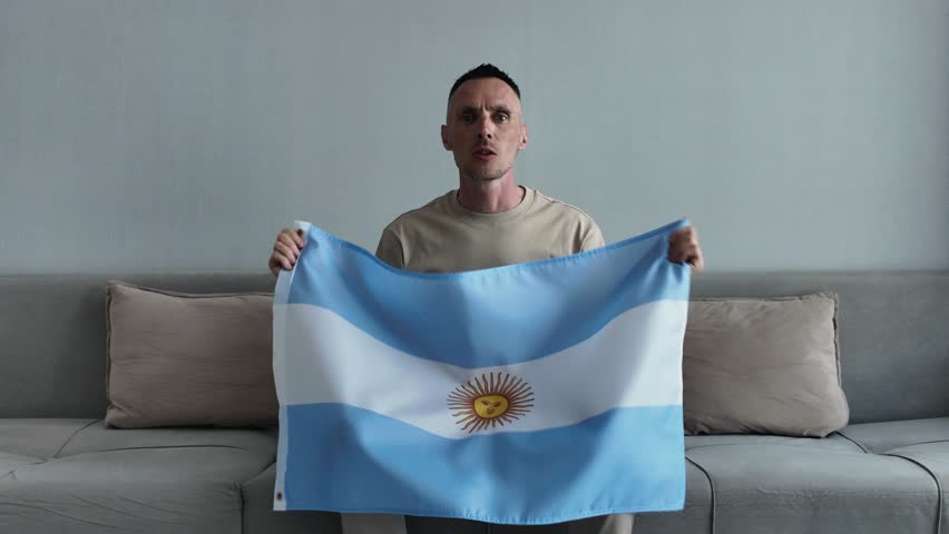 An enthusiastic Argentine sports fan holding the Argentine national flag shouts with joy and celebrates a goal while watching a football match at home