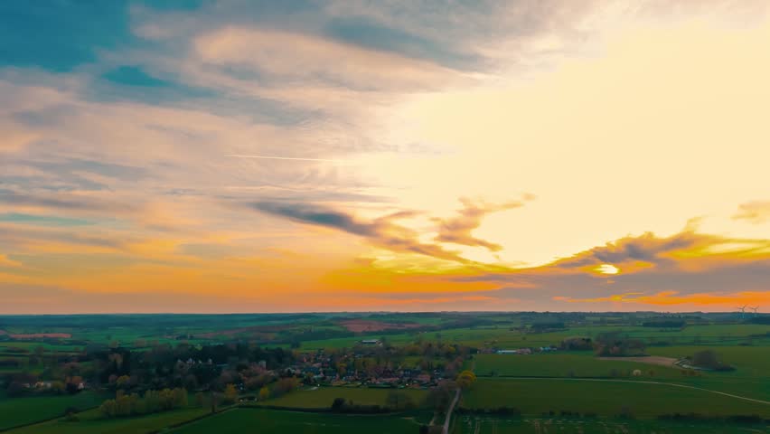 Timelapse agriculture field transitioning day to night across countryside panorama. Sunset over green farmland village horizon, dramatic sky clouds glowing orange blue evening light. Scenic rural