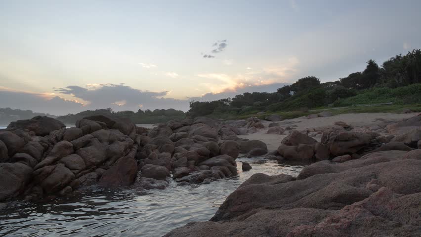 A tranquil scene unfolds as the sun begins to set over a lush, rugged coastline, casting a soft glow on the water and rocks along the South Coast, Kwa-Zulu Natal, South Africa.