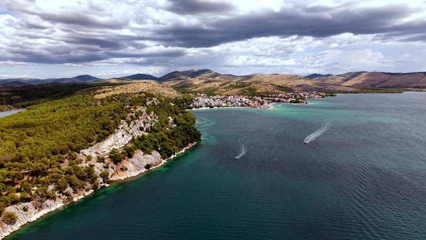 Aerial view of the scenic Adriatic coast in Croatia featuring a coastal town, rocky cliffs, and turquoise sea under a dramatic cloudy sky