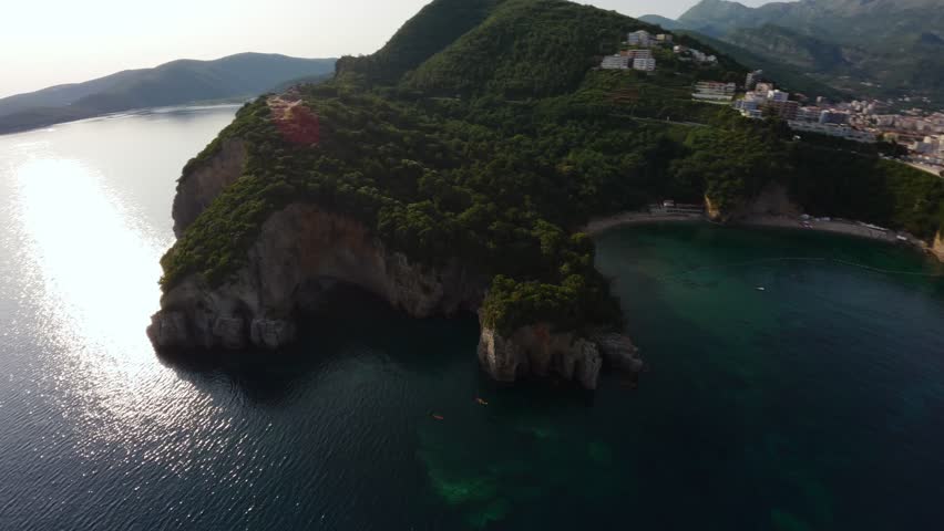 Aerial drone footage of Montenegro coastline bay at sunset with turquoise water, lush green hills, scenic shoreline and small houses, peaceful summer landscape with warm light and calm sea.