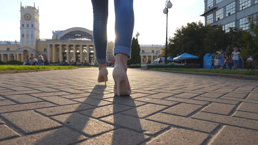 Camera follow to female legs in high heels shoes walking in urban street. Feet of young woman in high-heeled footwear going in city. Girl stepping on sidewalk with sun flare at background. Back view
