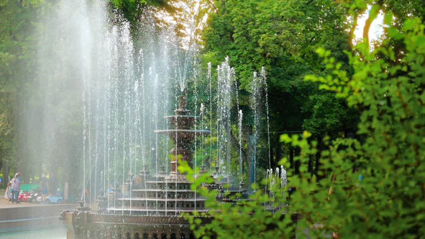 Ornate tiered fountain sprays water in lush green park on sunny day, people walk by.
