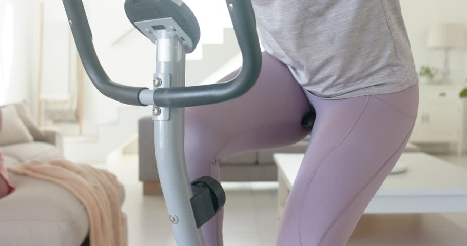 Female in purple leggings pedaling bike at home, starting workout, gripping bars checking console. Cardio, cycling, stationary, livingroom, daylight, minimalist, neutral