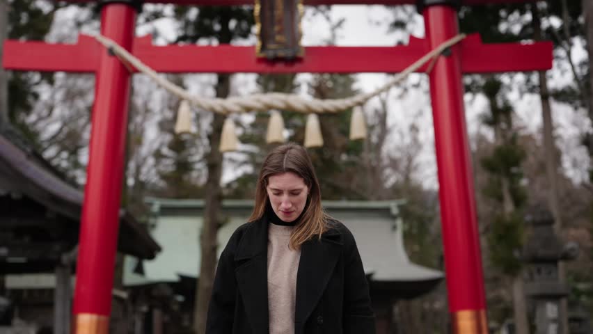 Woman standing and smiling under a traditional Shinto torii gate with sacred shimenawa rope at a Japanese shrine