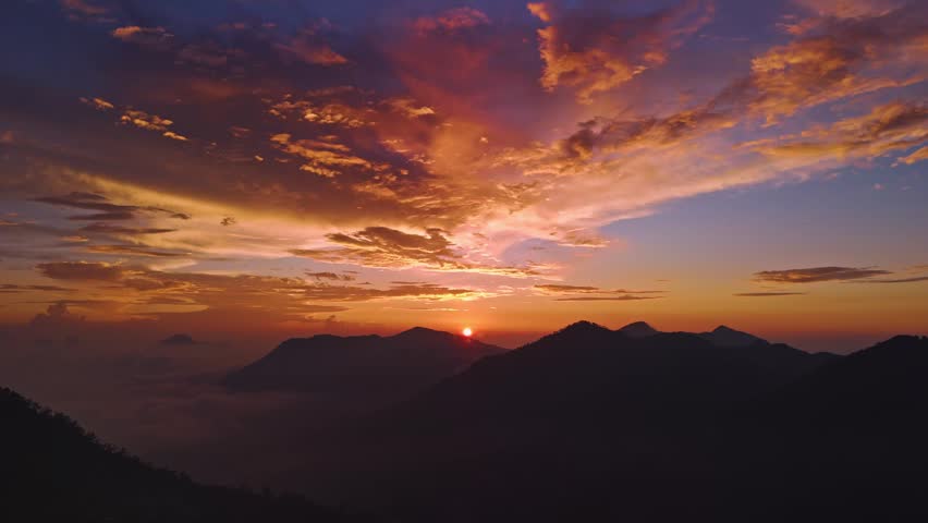 Sunset over mountain range with dramatic orange clouds. Sky glows with warm hues above silhouetted peaks. Mist drifts through valleys below ridges.