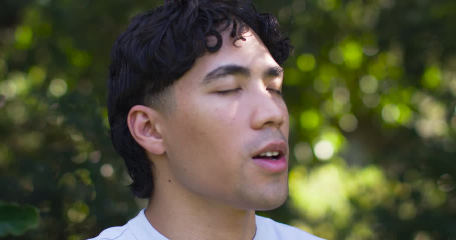 Asian male breathing, opening eyes, sensing dappled sunlight, wearing light shirt in park. Outdoors, nature, headshot, gaze, contemplation, greenery, bokeh
