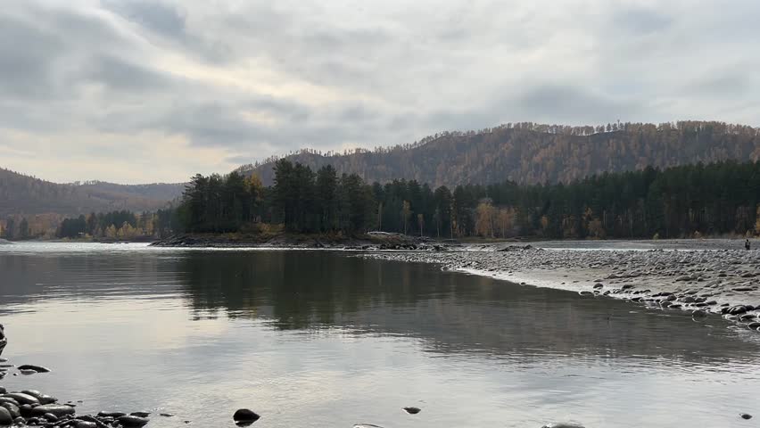 Serene wide river flows gently past a forest-covered island and a stony shore, with rolling hills in the background under a cloudy autumn sky.
