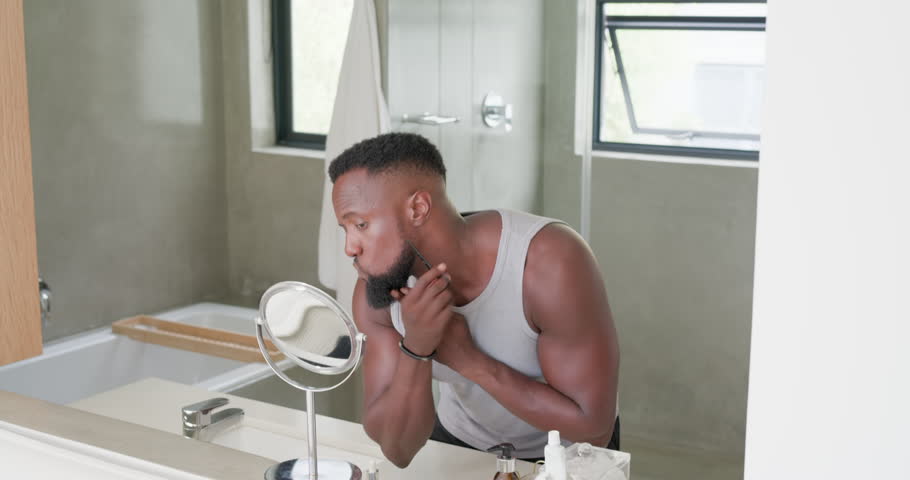 African American man seeing beard, trimming with trimmer at bathroom sink, applying oil to groom. Mirror, vanity, serum, dropper, razor, bathtub, towel