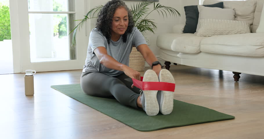 African American middle-aged woman pulling red looped band around shoes, toning back on green mat. Female, solo, minimalist, fitness, livingroom, seated, rowing
