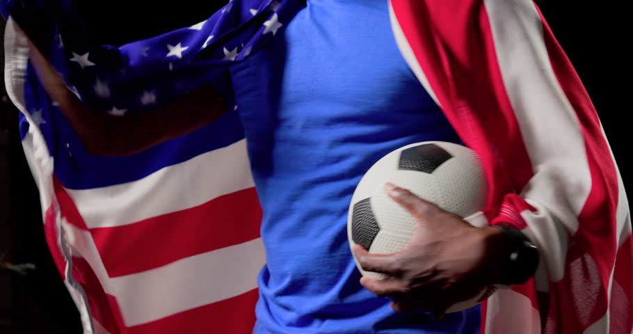 Adult African American male celebrating win, draping American flag, holding soccer ball in studio. Patriotic, cape, athlete, celebration, triumph, confidence, spotlight