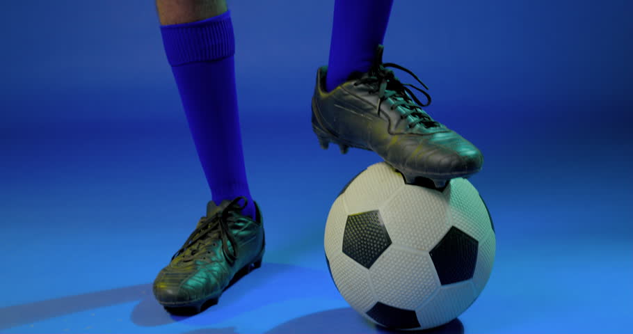 Man balancing soccer ball, shifting stance, crossing arms, posing in yellow jersey as camera rising. Athlete, studio, blue, shorts, socks, cleats, kit