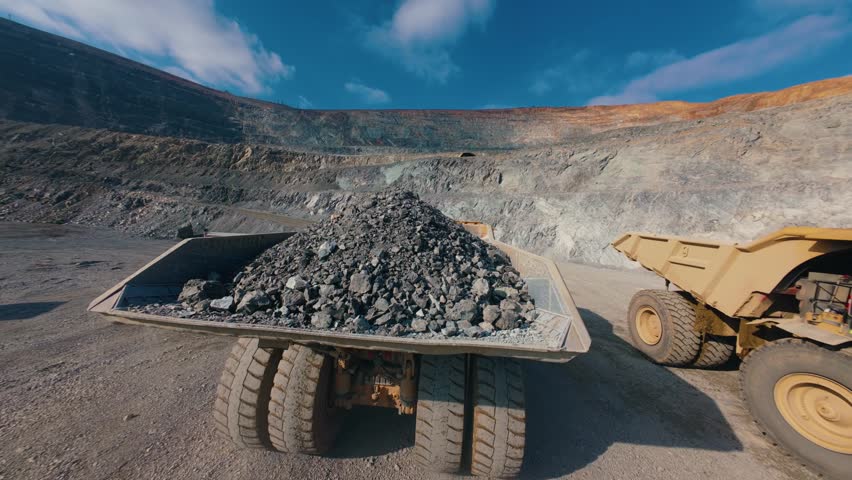 Aerial FPV heavy dump truck carrying load of gold ore inside open pit mine at midday, mining industry.