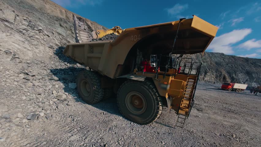 Tracking shot excavator loading gold ore to dump truck at open pit mine during daytime, aerial fpv.