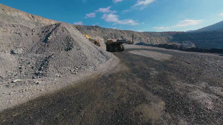 Aerial fpv view truck with gold ore while excavator loads in open pit mine quarry site during daytime.