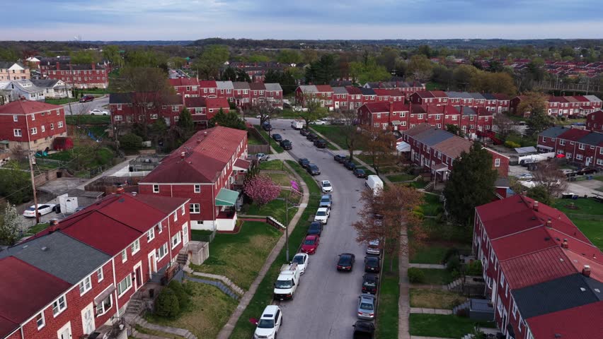Aerial view of a typical Baltimore, Maryland neighborhood with red brick townhouses, parked cars along residential streets and calm suburban atmosphere during spring. Establishing wide shot.