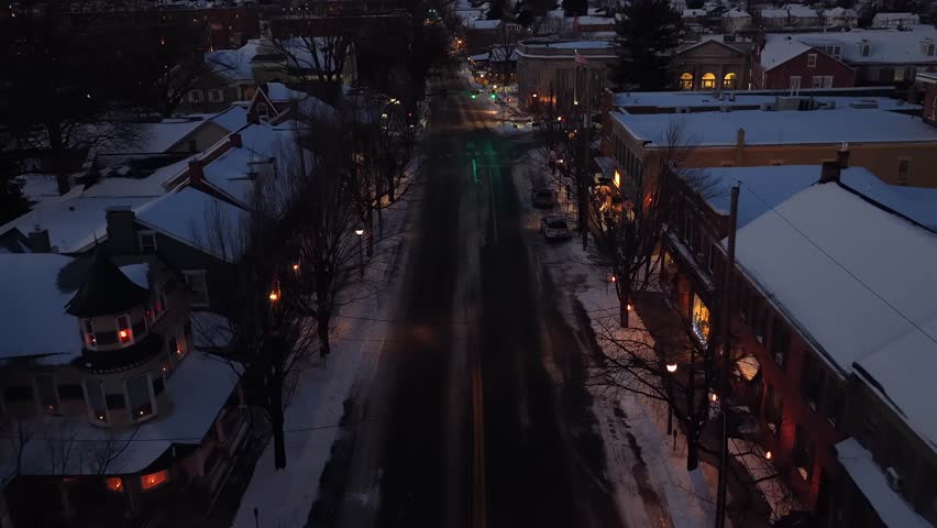 Aerial dusk view over Lititz, Pennsylvania, showing snow-covered colonial homes and quiet street illuminated by warm lantern lights. Peaceful winter evening scene. Flyover shot. Quiet night in USA.