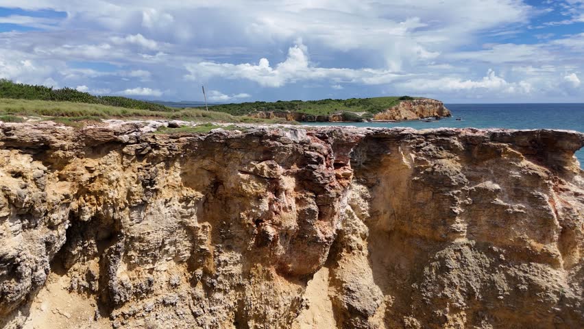 Aerial drone footage of rocky coastal cliffs and turquoise ocean in Cabo Rojo, Puerto Rico. Scenic tropical shoreline with rugged terrain, clear water, and dramatic clouds in the sky.