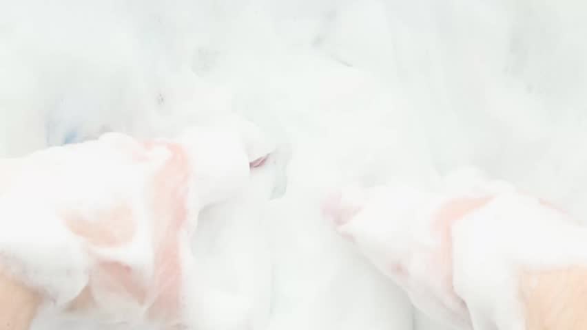 Close-up hands gently squeeze and lift dense white foam, slowly kneading airy suds back and forth until the frame fills with soft bubbles in a calming sensory self-care moment.
