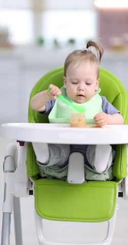 Cute little child eating baby food on feeding chair at home