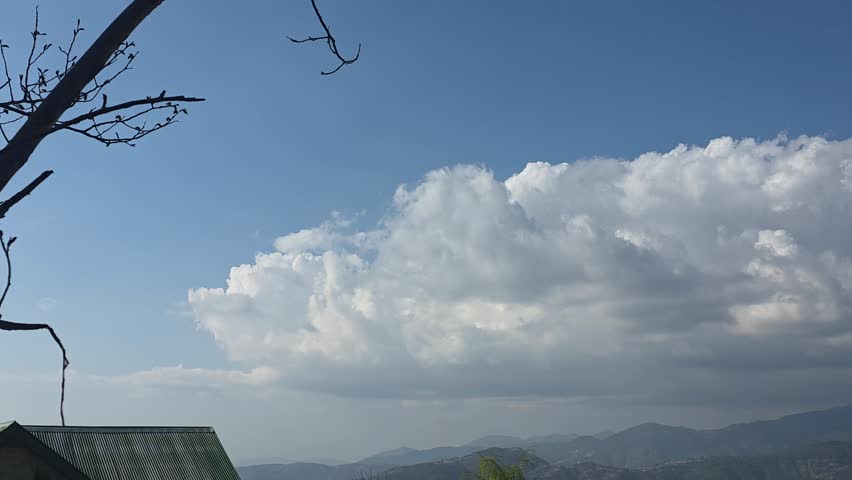  Right Pan of Fluffy White Cumulus Clouds Over Hilly Landscape. A smooth horizontal camera pan from left to right, capturing a bright blue sky filled with massive, towering white clouds. The foreground shows a silhouette of mountains and a small building with power lines, creating a scenic and vast atmosphere of a suburban hill station.