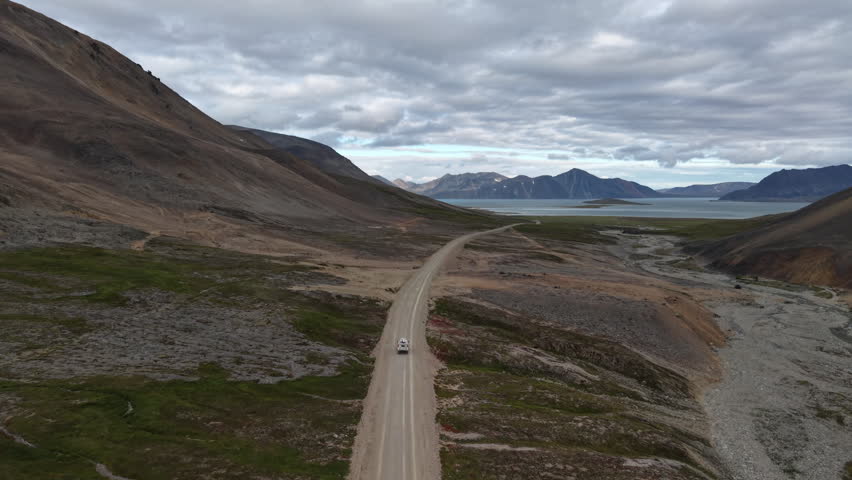Drone flyover tracks right along a remote tundra road toward the coastal mountains and bay, with a lone vehicle moving ahead under dramatic cloud cover. Russia, Chukotka, Provideniya.