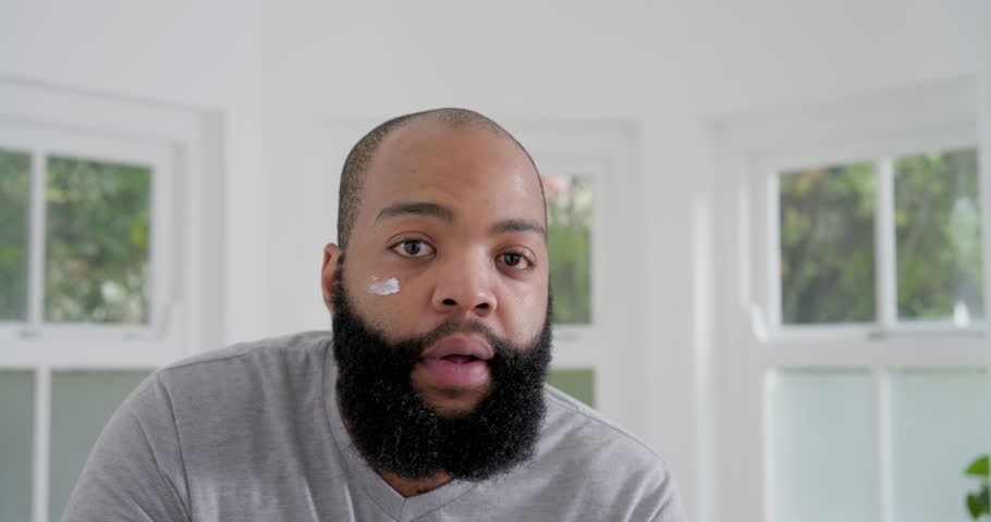 African man in gray shirt at home by windows starting from cheek-cream-dot, dabbing for skincare. Beard, portrait, model, windows, interior, bright, modern