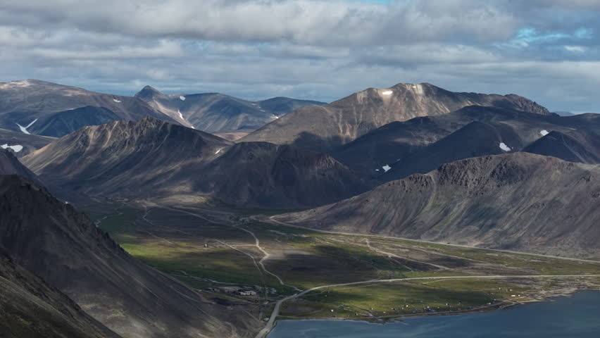 Overcast daylight over remote mountains and green valley near Provideniya, Chukotka. Drone hovers above winding road and river plain, capturing vast terrain. aerial establishing shot