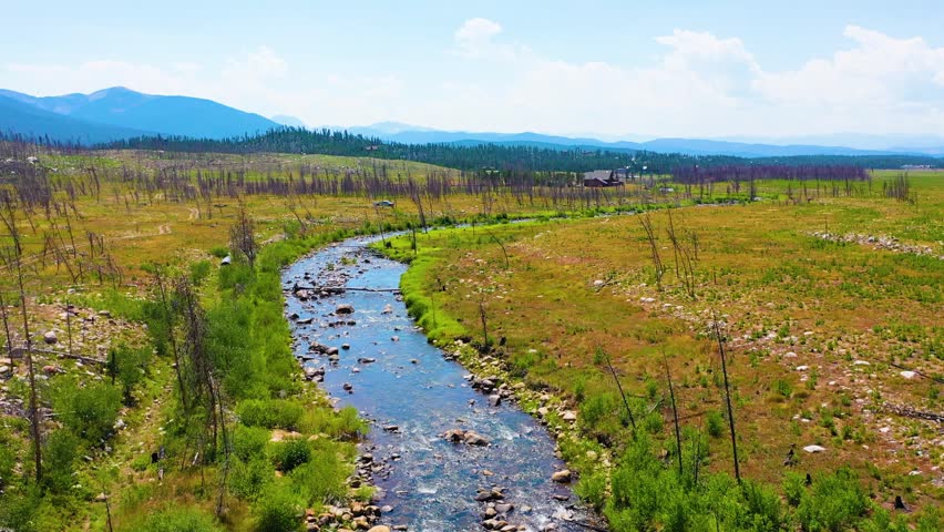Drone POV pulling back over an Alaska style wilderness creek through a broad valley with rocky shallows vivid green banks sparse timber and distant mountain layers under summer clouds