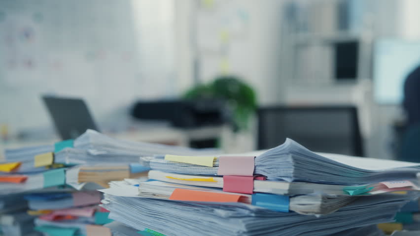 Closeup overworked young Latin businessman reading document in large stack of paperwork file at office desk. Tax audit deadline, accounting stress, corporate burnout, bookkeeping, bureaucracy concept.