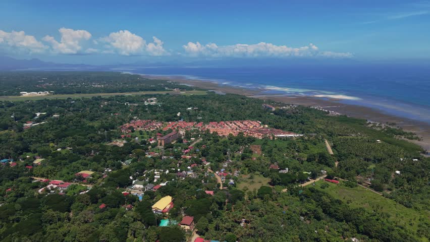 Forward aerial flyover above Bankao-Bankao in Puerto Princesa, Palawan, Philippines, with the drone moving steadily ahead at moderate altitude. The frame opens onto a quiet coastal town with rows of small houses and bright rooftops surrounded by dense green vegetation. Low palm canopies and clusters of trees fill the spaces between the buildings, blending the settlement into the surrounding tropical landscape. To the right of the frame, a wide stretch of tidal flats extends along the shore, where shallow water and exposed reef textures shape the coastline at low tide. Far in the background, layered mountains rise across the horizon under a bright sky filled with soft cumulus clouds. The deep blue of the open sea curves around the headland, separating the inhabited shore from the wider bay beyond. Daylight falls evenly across the rooftops and trees, bringing out warm earth tones, soft greens, and cool ocean blues in a calm balanced palette. The slow forward movement gives a clear sense of scale and direction as more of the coastal community and its natural surroundings come into view. The footage offers a grounded look at everyday tropical life along the shoreline of Palawan in the Philippines.