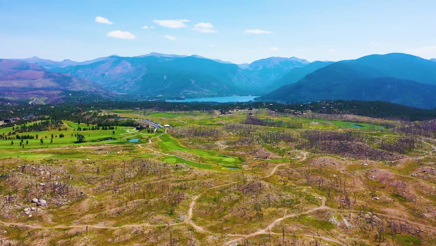 Cinematic aerial of a Yellowstone style mountain valley in Wyoming with open meadowland a distant lake rolling terrain forest bands and dramatic peaks under bright summer skies