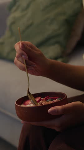 Vertical side view slowmo shot of young Black woman eating delicious yogurt bowl with granola and strawberries, having healthy breakfast high in fiber and protein