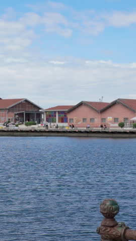 A vertical panoramic view of the historic port area in Thessaloniki, Greece, featuring redroofed warehouses, people walking on the pier, and industrial cranes under a blue sky.