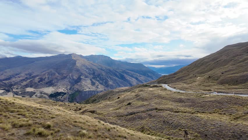 4K 30fps cinematic high-angle view of a vast, majestic mountain valley on the road to Queenstown, South Island, New Zealand. The footage captures the epic scale of the alpine landscape with winding roads, golden tussock grass, and rugged mountain ranges under a clear blue sky. This wide panoramic shot shows the pristine wilderness of the Otago region, typical of the Lindis Pass or Crown Range scenery. Perfect for travel documentaries, car commercials, adventure tourism, and vanlife lifestyle content. The high-altitude perspective emphasizes the solitude and breathtaking natural beauty of the New Zealand Alps during a bright sunny day.