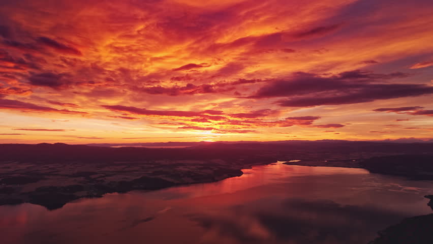 Aerial view of a dramatic fiery sunset over a fjord with intense red clouds and reflections on water. Cinematic wide landscape with mountains and vibrant sky in northern Europe.