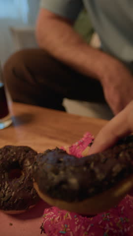 Vertical POV close up shot of bearded young Caucasian man biting delicious chocolate donut, enjoying pastry at home in evening